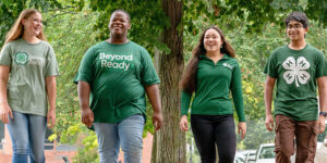 Four individuals wearing green 4-H shirts walk together outdoors under a large tree. The shirts feature 4-H clover logos and slogans such as “Beyond Ready.” This image represents teamwork and community involvement for One Day service activities.