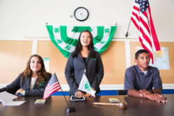Three people at a table with 4-H and U.S. flags and a green banner on the wall. Three people at a table with 4-H and U.S. flags and a green banner on the wall.