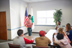 Person speaking to a group seated at a table in a room with U.S. and 4-H flags. Person speaking to a group seated at a table in a room with U.S. and 4-H flags.