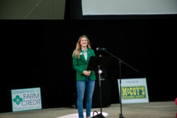 Person in a green blazer presenting on stage near a microphone stand and sponsor signs. Person in a green blazer presenting on stage near a microphone stand and sponsor signs.