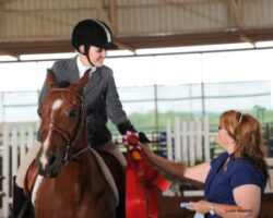 A rider on horseback accepts a red ribbon from an official inside an indoor arena during an equestrian competition. A rider on horseback accepts a red ribbon from an official inside an indoor arena during an equestrian competition.