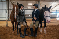 Two riders in formal English riding attire stand inside an indoor arena holding their horses by the reins. Two riders in formal English riding attire stand inside an indoor arena holding their horses by the reins.
