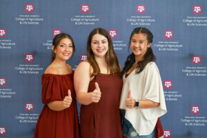 Three individuals standing in front of a Texas A&M University College of Agriculture & Life Sciences backdrop at Weekend in Aggieland, each giving a thumbs-up gesture. They are dressed in maroon and white attire, representing school spirit.