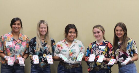Five people in floral shirts hold decorated white fabric pieces indoors, likely during a craft event.