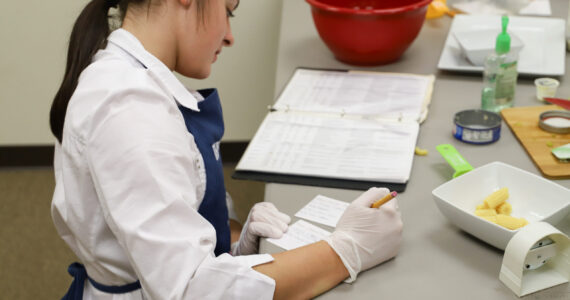 Individual wearing gloves and apron writing notes in a recipe binder with food ingredients and utensils on the table.