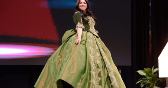 Individual wearing an elaborate green gown with lace details while walking on a stage during a fashion event.