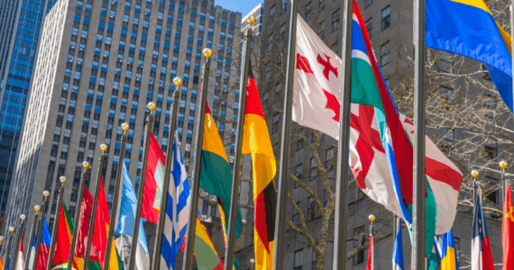 Rows of international flags displayed on tall flagpoles in front of high-rise buildings.