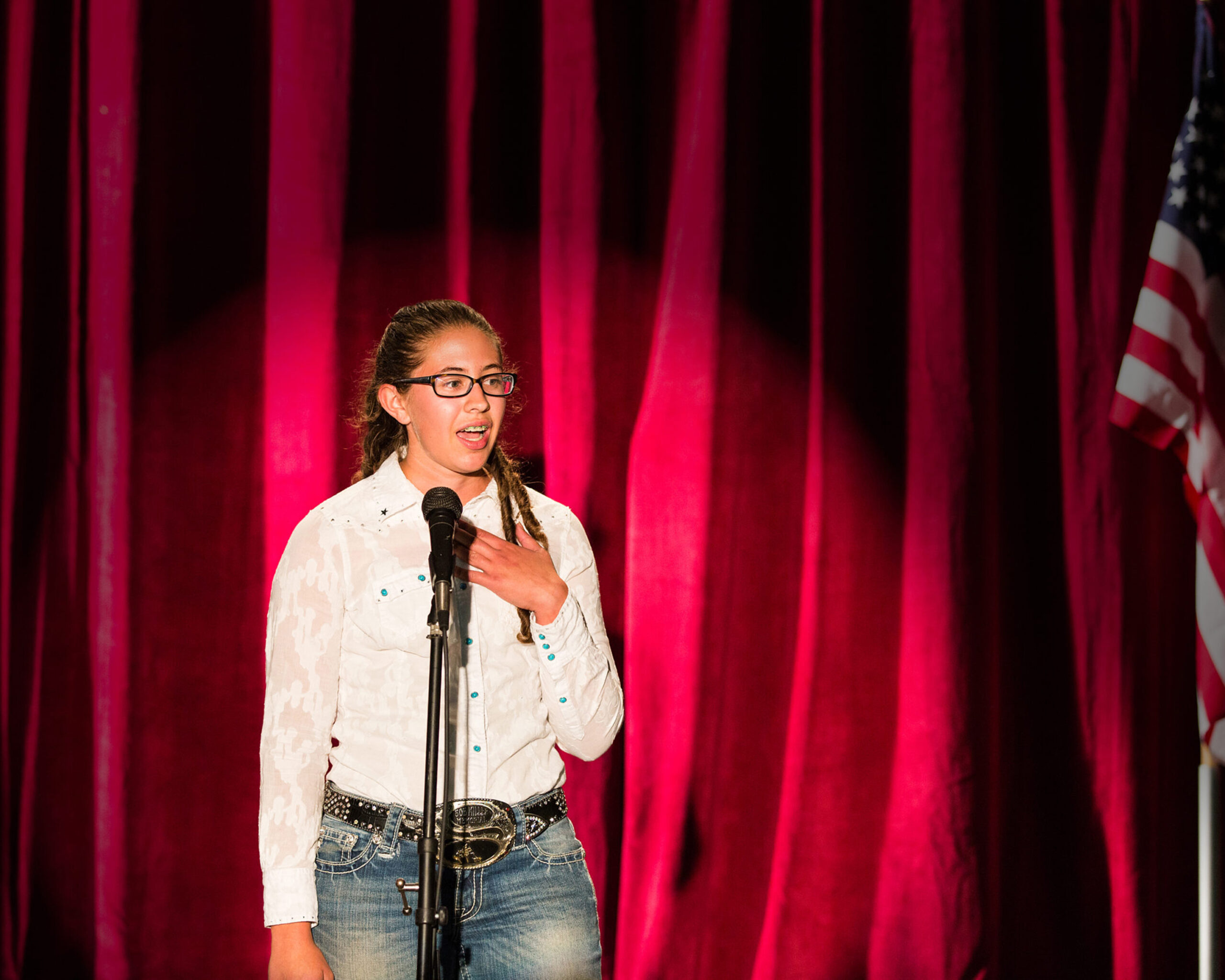 Presenting a speech at a microphone during a public speaking contest. Individual standing at a microphone on stage with a red curtain backdrop and U.S. flag.