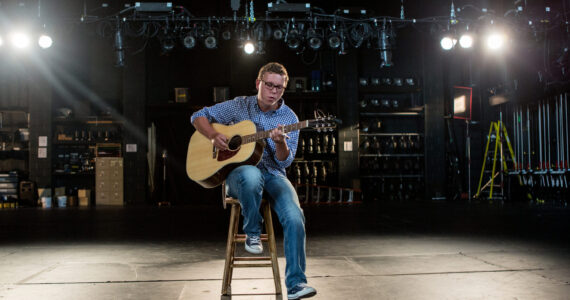 Musician playing an acoustic guitar under stage lights in an empty theater.